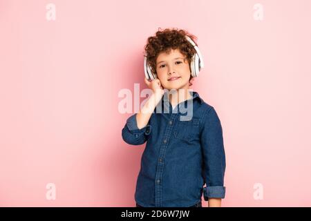 pleased boy listening music and touching wireless headphones on pink Stock Photo