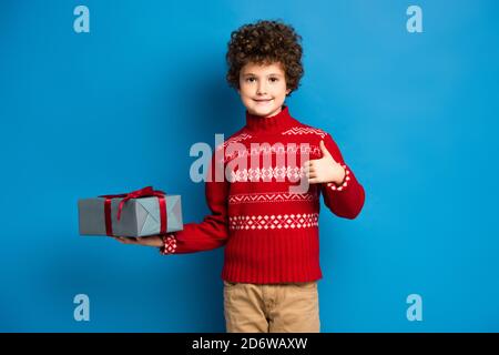 curly boy in red sweater with ornament holding present and showing thumb up on blue Stock Photo