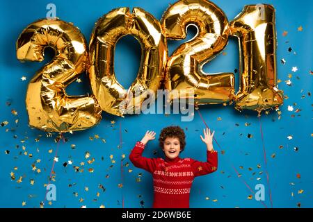 excited boy in red sweater gesturing near golden balloons with 2021 numbers and confetti on blue Stock Photo