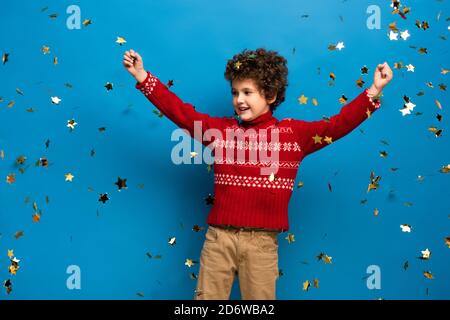 excited boy with hands above head in red sweater near golden confetti on blue Stock Photo