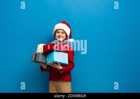 happy boy in santa hat and sweater holding presents on blue Stock Photo