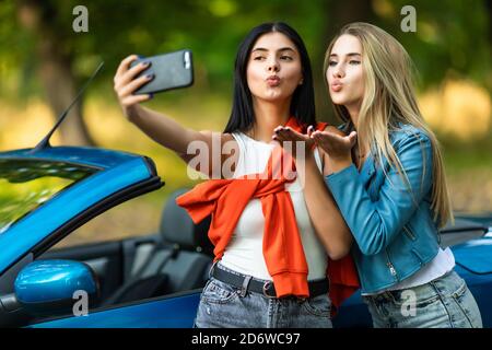 Young pretty two women blow kiss taking a selfie while standing near convertible car on the street Stock Photo