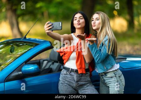 Young pretty two women blow kiss taking a selfie while standing near convertible car on the street Stock Photo