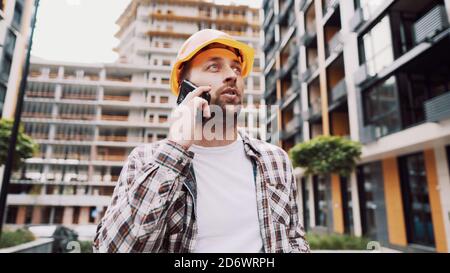 Portrait of young handyman making call while standing at construction area. Engineer talking on the phone on a construction site. Builder in helmet Stock Photo