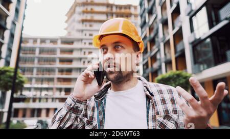 Portrait of young handyman making call while standing at construction area. Engineer talking on the phone on a construction site. Builder in helmet Stock Photo