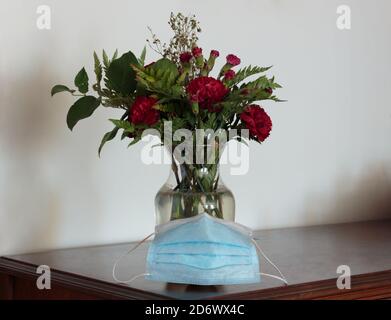 a blue face mask set in front of a vase of red carnation flowers on a dark wooden table next to a white wall with copy space Stock Photo