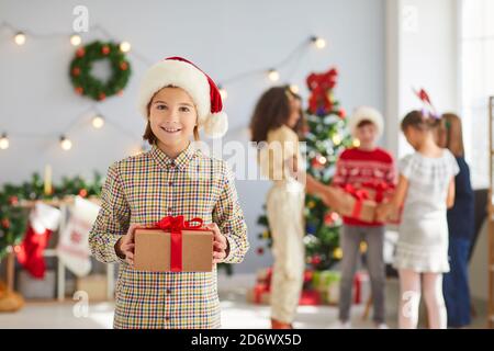 Happy little boy in a red Santa Claus hat holds a gift box wrapped in kraft paper and red ribbon.
