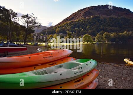 A stack of canoes at Glenridding Sailing Centre line the shore of Ullswater in the Lake District.  The Inn On The Lake can be seen behind the trees. Stock Photo