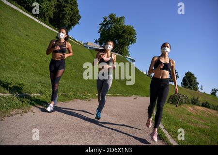Three multiethnic women group wear face masks run keep social distance outdoor. Stock Photo