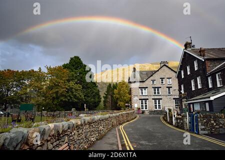 A rainbow above the Glenridding Hotel on the edge of Ullswater in the Lake District National Park.  A stone wall lines the road leading to the hotel. Stock Photo
