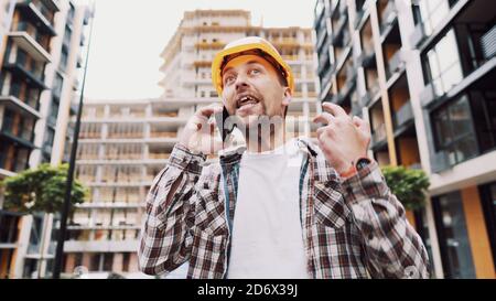 Angry builder, dissatisfied with deadline of work, swears at stress while talking to foreman on phone background of construction site. Wrathful Stock Photo