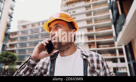 Angry male architect in orange hard hat talks swears on phone at construction site. Builder in plaid shirt emotionally displeased negotiates by cell Stock Photo