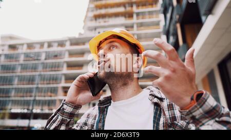 Angry male architect in orange hard hat talks swears on phone at construction site. Builder in plaid shirt emotionally displeased negotiates by cell Stock Photo