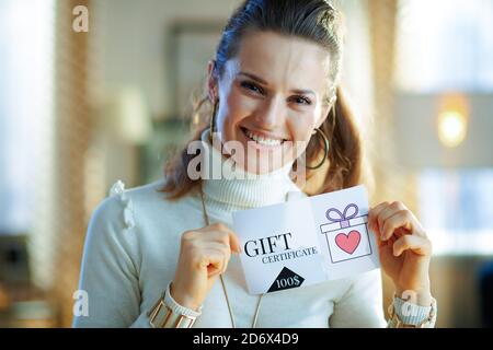 smiling middle age woman in white sweater and skirt in the modern house in sunny winter day showing gift certificate.