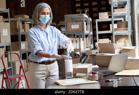 Older female entrepreneur wearing mask using sanitizer working in warehouse. Stock Photo