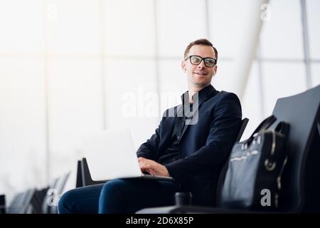 Man with suitcase sitting in airport waiting area while listening music using airpods Stock Photo