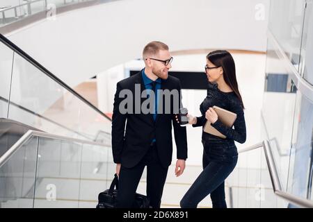 Beautiful business woman and man their holding a tablet in hands and talking to each other while standing at staircase at modern office building. Stock Photo