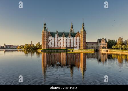 The Royal Frederiksborg castle glimmers in the sunshine and is reflected in the lake with a swimming swan, Hillerod, Denmark, October 17, 2020