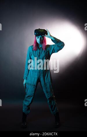 Young woman with pink hair and in a jumpsuit uses virtual reality glasses, is in the game, black background. VR simulation. Stock Photo