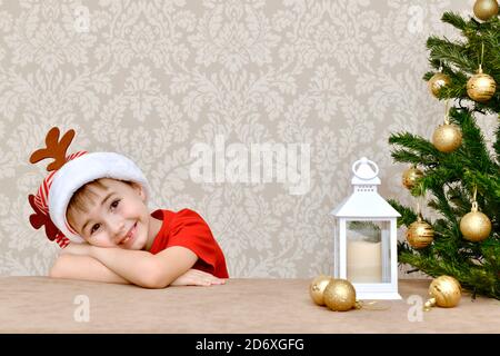 A gentle smiling kid without a tooth lay on folded handles near the Christmas tree. In a carnival cap with deer horns. Stock Photo