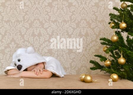 A child in a striped cap with deer horns laughs sitting at the table at the artificial Christmas tree and white lantern. Stock Photo