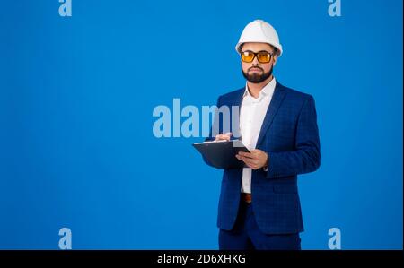 A young professional wearing a hard hat and carrying a clipboard on blue background Stock Photo