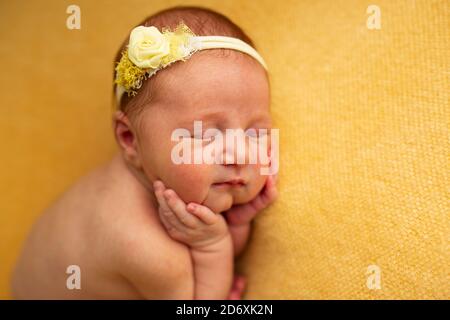 portrait of a beautiful nine-day-old baby girl sleeping in a curled up fetal position on a yellow blanket. She is wearing a yellow rose headband Stock Photo