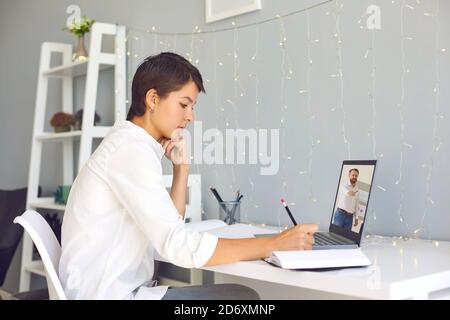 Young woman sitting at desk at home, watching educational business video on laptop and taking notes Stock Photo