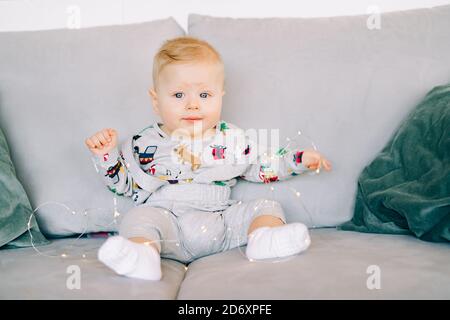 a small cute, cheerful boy sits in a festive Christmas costume on a gray sofa and plays with a bright light garland.Christmas concept. Stock Photo