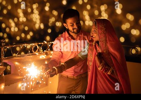 Happy young indian couple celebrating diwali festival both holding sparkler in hand and wearing traditional wear or ethnic outfit on auspicious evenin Stock Photo