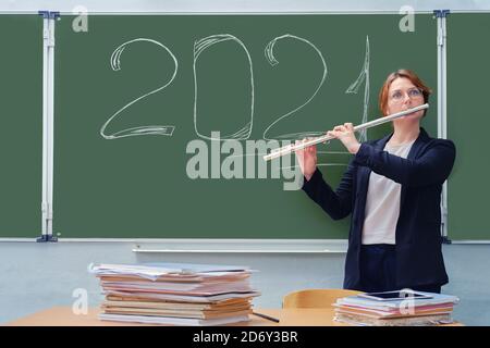 A music teacher plays a musical instrument on the background of a blackboard with the text 2021. Learning to play wind instruments in quarantine durin Stock Photo