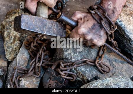 The dirty hands of a slave holding a hammer are entangled in a rusty chain with a large padlock on a background of stones. Stock Photo