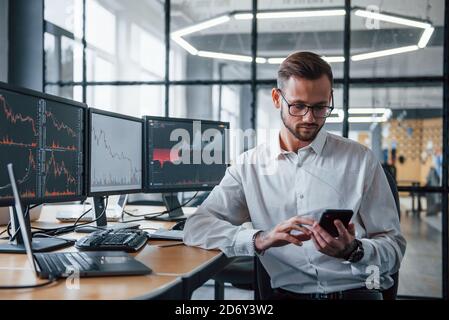 With phone in hands. Male stockbroker in formal clothes works in the office with financial market Stock Photo