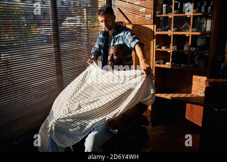 Bearded barber putting hairdressing cape on client at barbershop Stock Photo