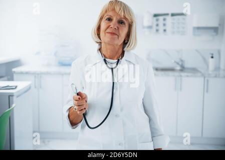 Holds stethoscope in hand. Portrait of senior doctor in white uniform that stands in the clinic Stock Photo