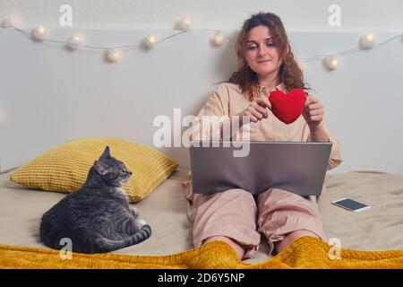 Happy woman is texting on online dating sites. Girl sitting with cat in home bedroom, internet love concept Stock Photo