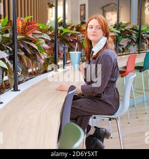 The woman removed the medical mask after the end of the coronavirus quarantine at the bar in a restaurant. Portrait of a red-haired girl in a cafe clo Stock Photo