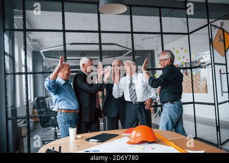 Job well done, gives high five to each other. Aged team of elderly businessman architects stands in the office together Stock Photo