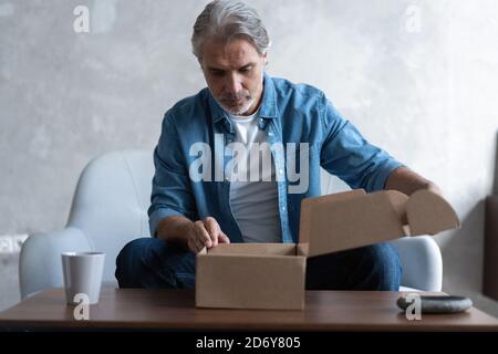 Smiling man consumer open cardboard box get postal parcel, male customer receive carton package sit on sofa at home Stock Photo