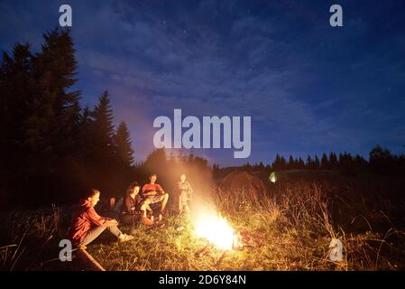 Group of friends sitting near campfire in high green grass, illuminated tent at night camp in the mountains, surrounded by spruce trees under magical Stock Photo