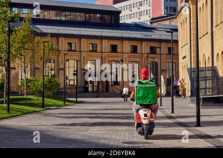 Rear view of courier with refrigerator bag riding scooter along beautiful sunny street with small houses. Food delivery concept Stock Photo