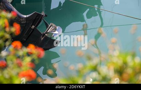 A silver boat propeller on a black motor framed by red and white flowers, raised out of the water, reflected on the turquoise waters of the Ionian Sea. Stock Photo