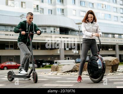 Young man riding electric kick scooter and young woman in casual wear riding unicycle on city street Stock Photo