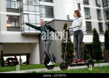 Young man riding unicycle and young woman in casual wear riding on electric kick scooter on city street Stock Photo