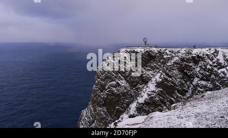 Popular globe sculpture on snow-covered rock cliff of North Cape (Nordkapp), Norway, Scandinavia with few tourists and rough arctic sea in winter. Stock Photo