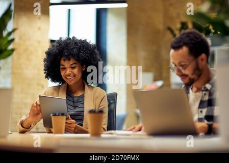 Two young happy multiracial business people sitting at the desk, using modern technologies while working together in coworking space. Office life, teamwork and business Stock Photo