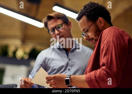Two focused diverse business colleagues analyzing document and discussing something while standing in coworking space, working together. Office life, teamwork and business concept Stock Photo