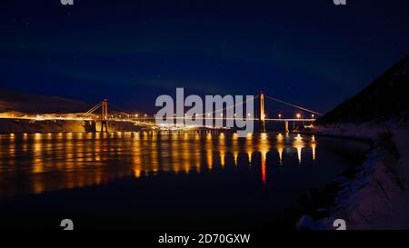 Illuminated Kvalsund suspension bridge near Hammerfest, Norway at night in winter time with slightly visible aurora and reflections in water. Stock Photo