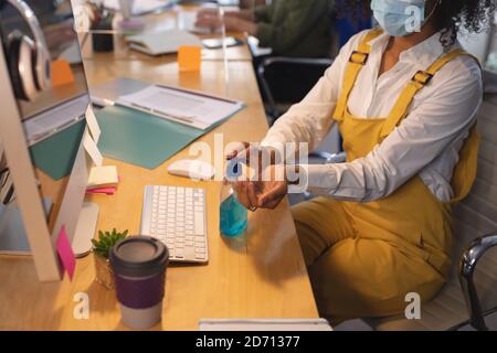 Mixed race female creative sitting at a desk in an office disinfecting hands with hand santiser. Health and hygiene in workplace during Coronavirus Co Stock Photo