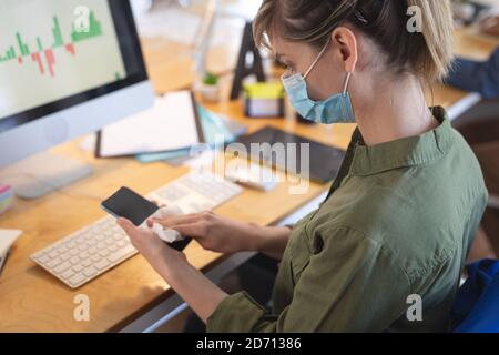 Caucasian female business creative sitting at desk in an office wearing face mask disinfecting her smartphone. Health and hygiene in workplace during Stock Photo